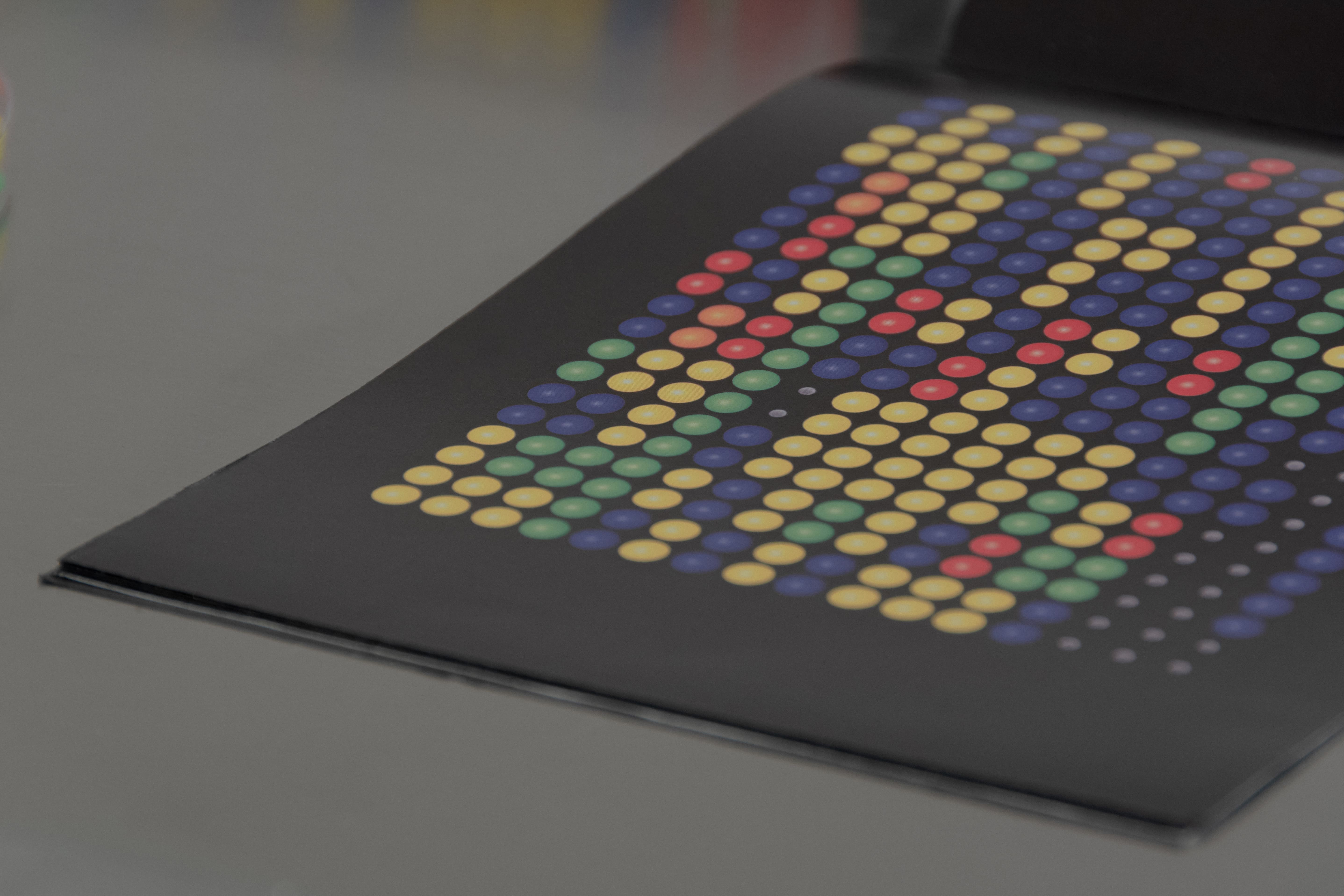 A black page is open in a book with a colorful pattern of evenly spaced, small dots in rows, featuring blue, yellow, red, and green hues. A blurred, neutral background creates a calm atmosphere.