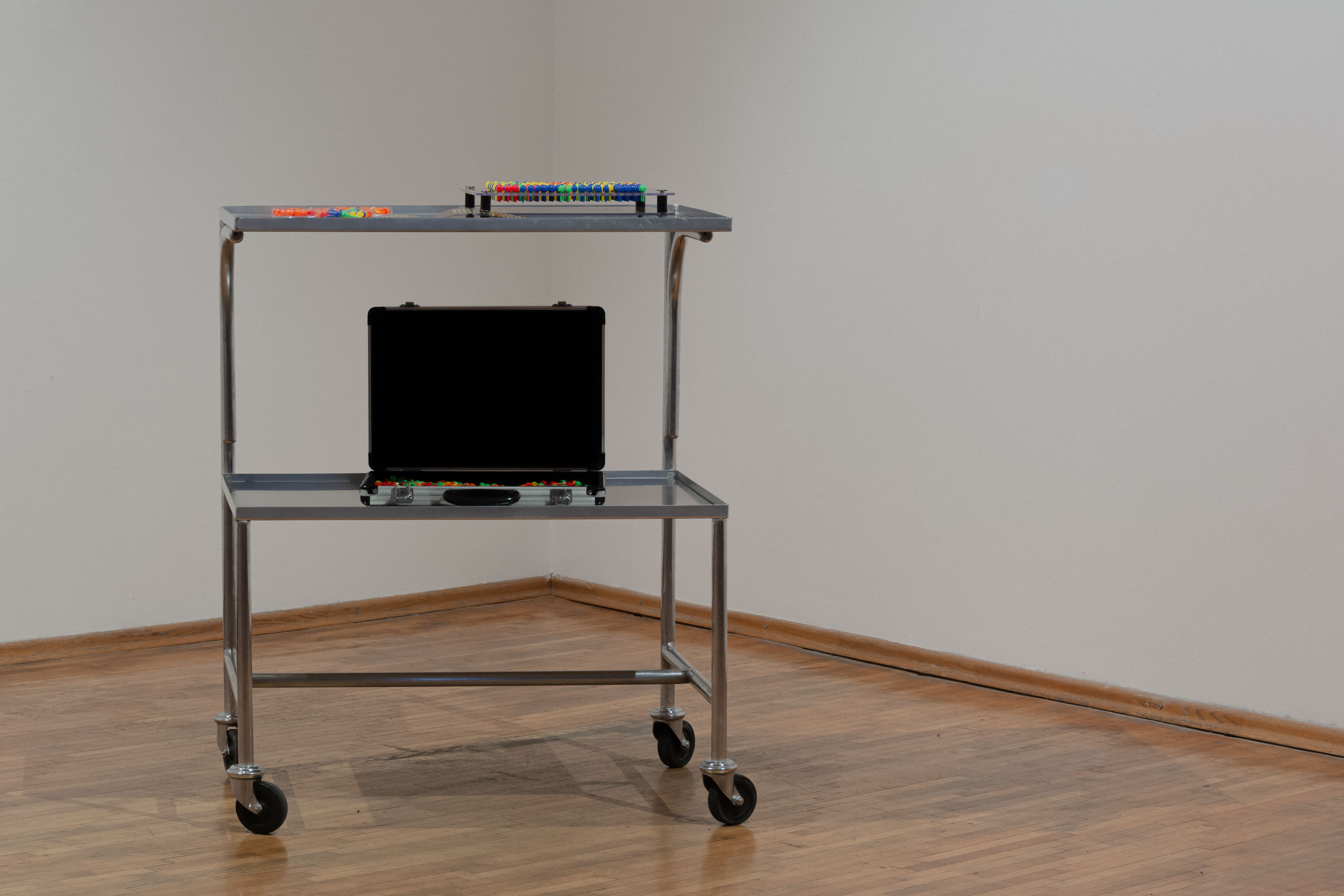 A metal trolley with two shelves on wheels, placed in a corner. The top shelf holds colorful objects, while the bottom shelf has a closed black briefcase.
