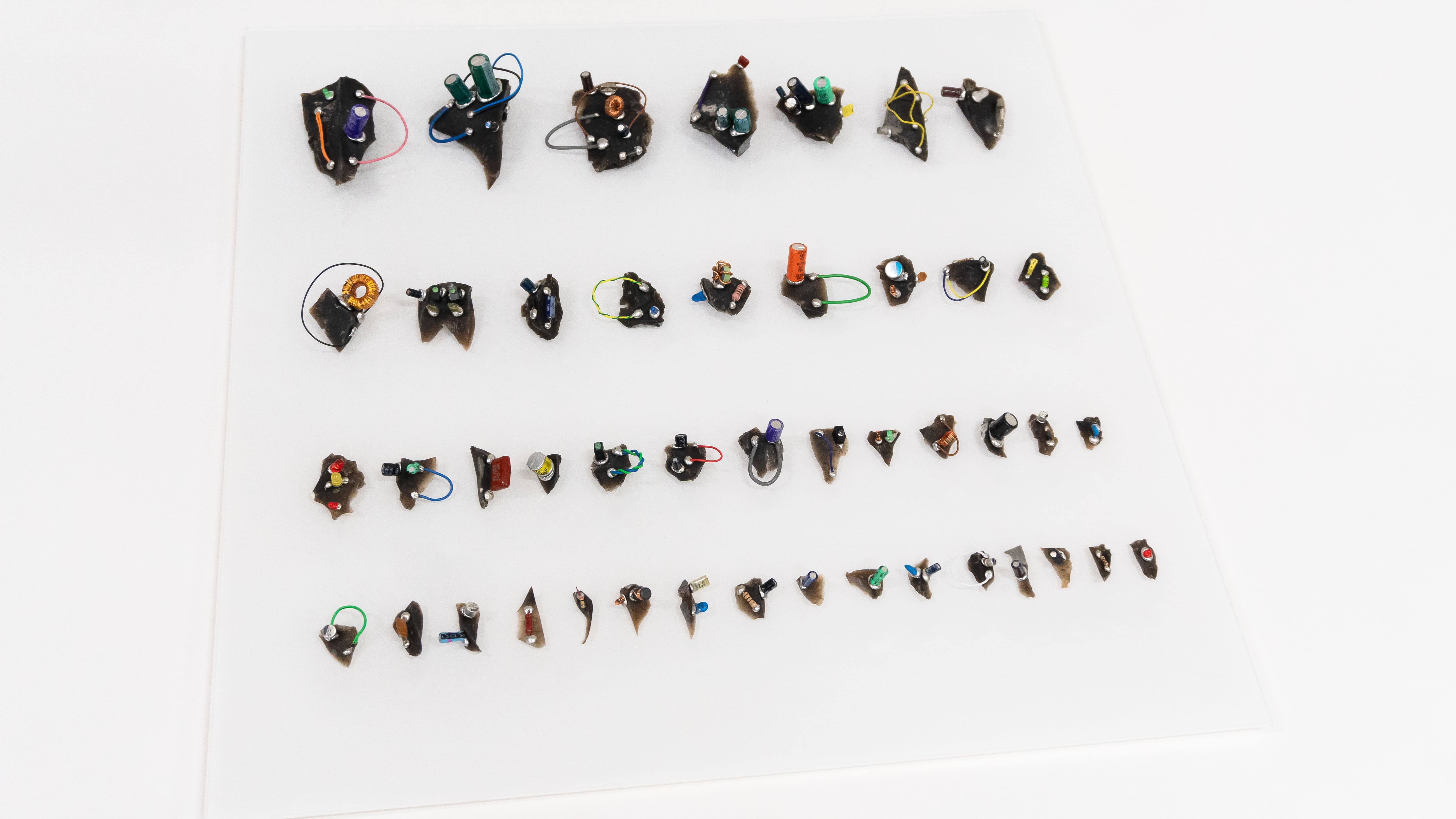 Array of small, abstract sculptures resembling circuit boards, with colorful wires and components, neatly aligned on a white background.