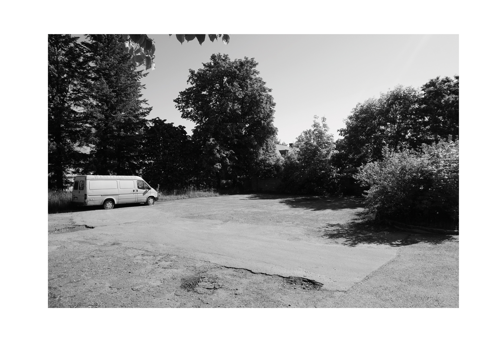 Black and white image of an empty parking lot with a single white van parked on the left. Surrounded by tall trees, creating a serene, quiet atmosphere.